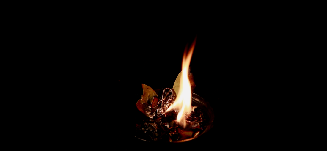 a plume of fire & herbs rosemary & bay laurel burning in a vessel, with a silver pendant in amongst the herbs & ashes